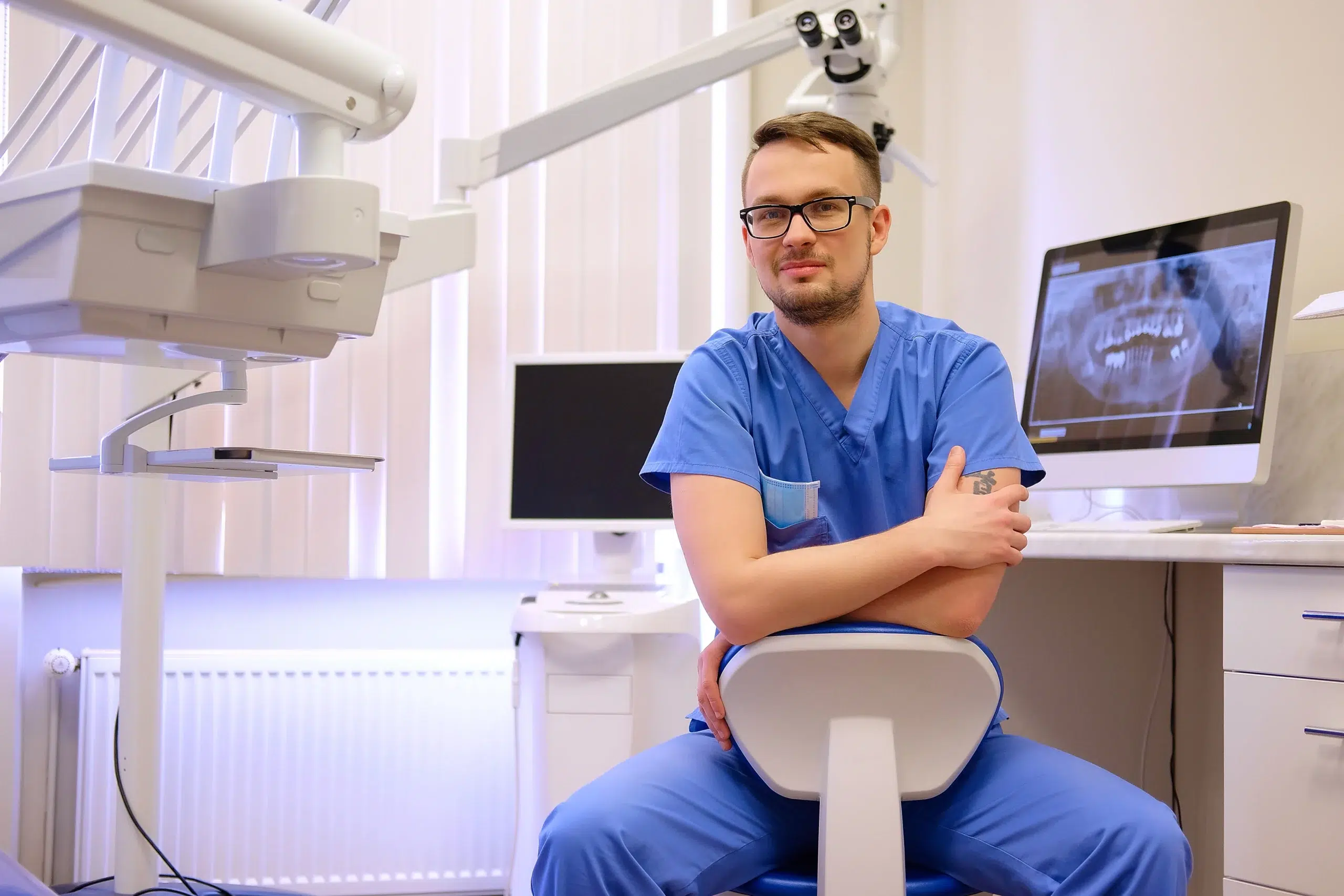 portrait handsome bearded dentist male glasses wearing blue uniform sitting dentist office looking camera scaled